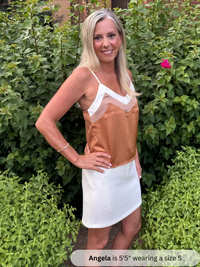 Woman wearing a brown and white dress standing in front of green bushes.