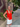 /Tomato Red/ in a red top and white skirt standing on a sidewalk with a stone building and trees in the background.