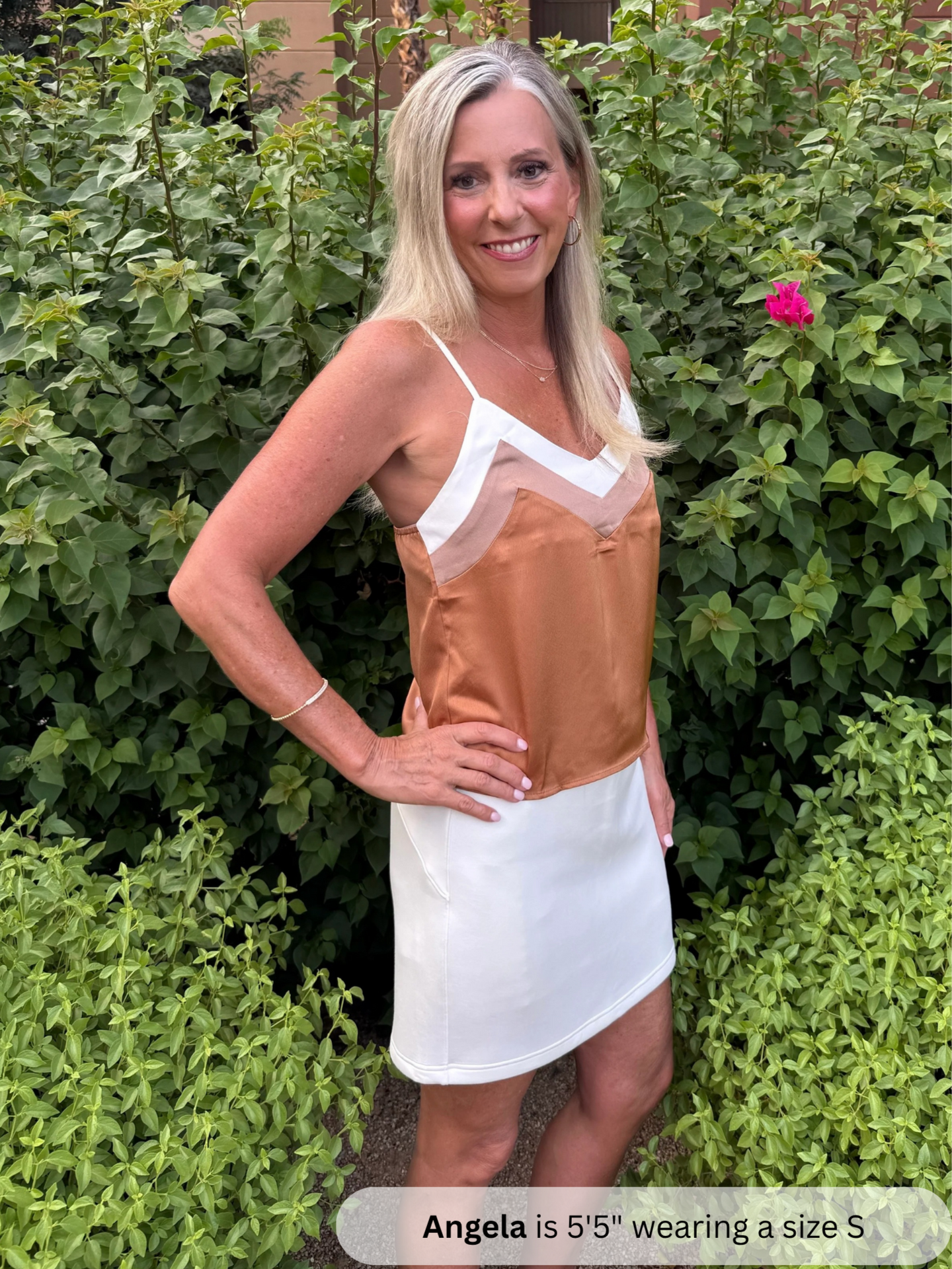 Woman wearing a brown and white dress standing in front of green bushes.