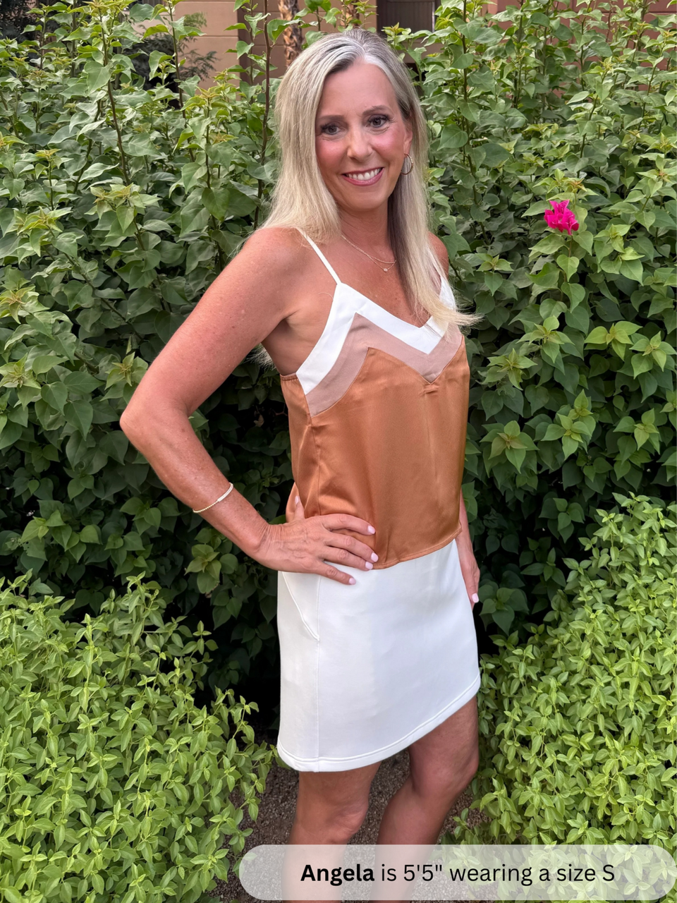 Woman wearing a brown and white dress standing in front of green bushes.
