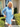 Woman wearing a light blue dress standing outside a house with 'Welcome' sign.