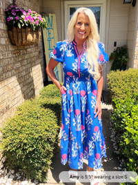 Woman in a blue floral dress standing outside a house with plants and flowers.