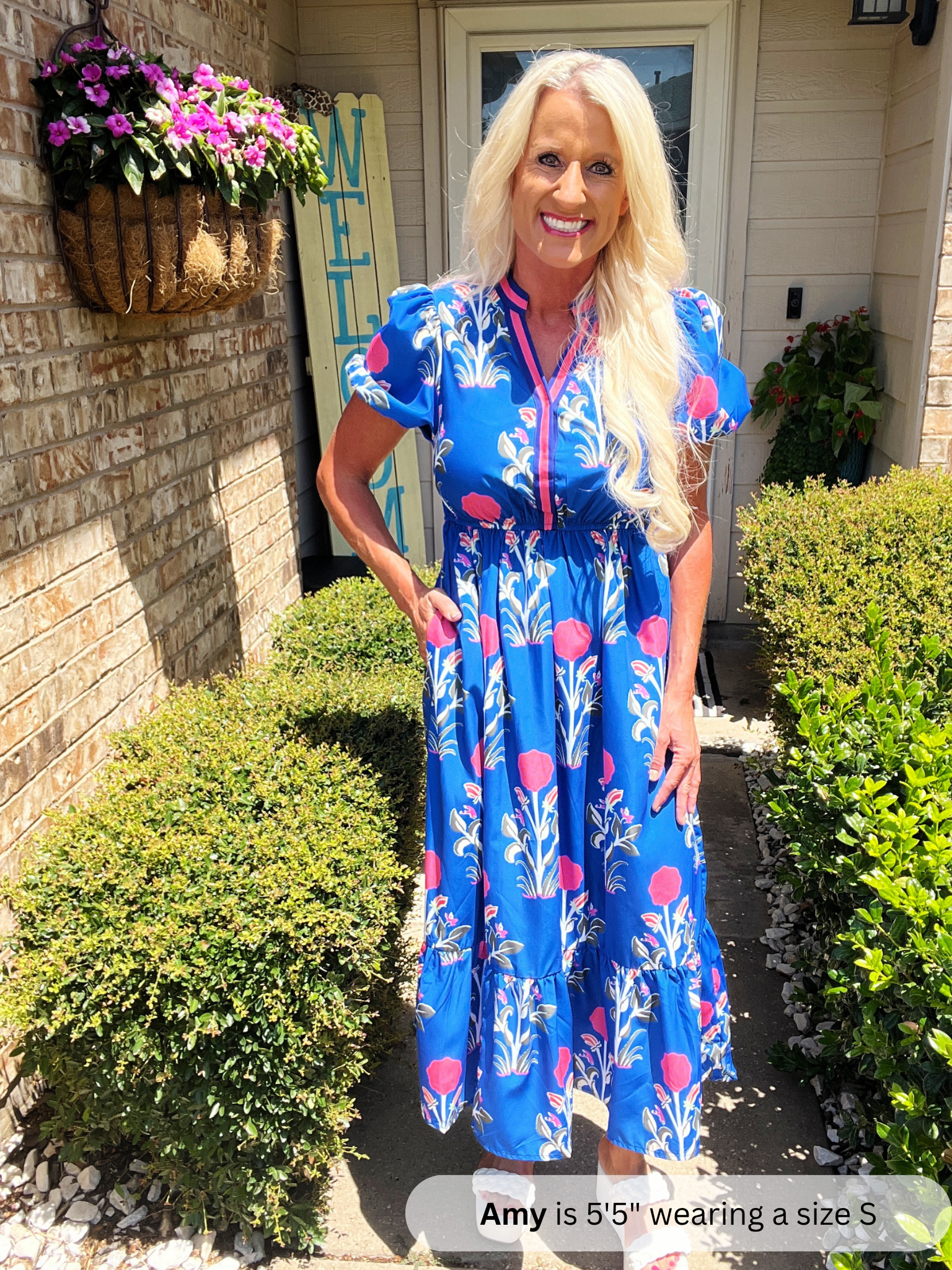 Woman in a blue floral dress standing outside a house with plants and flowers.