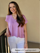 /Mauve/ Woman wearing a purple shirt and white pants standing against a beige wall.