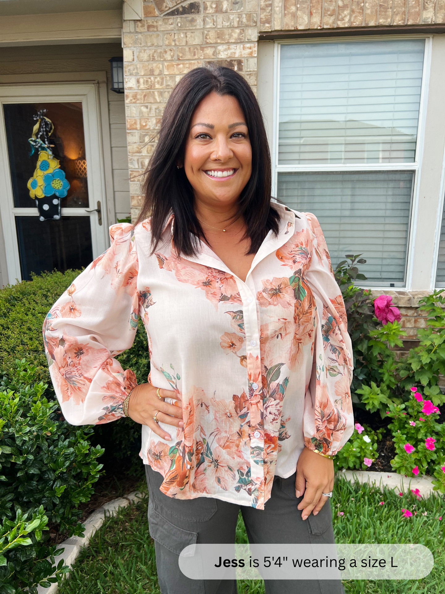 Woman wearing a floral blouse standing outside a house with flowers and a door decoration.