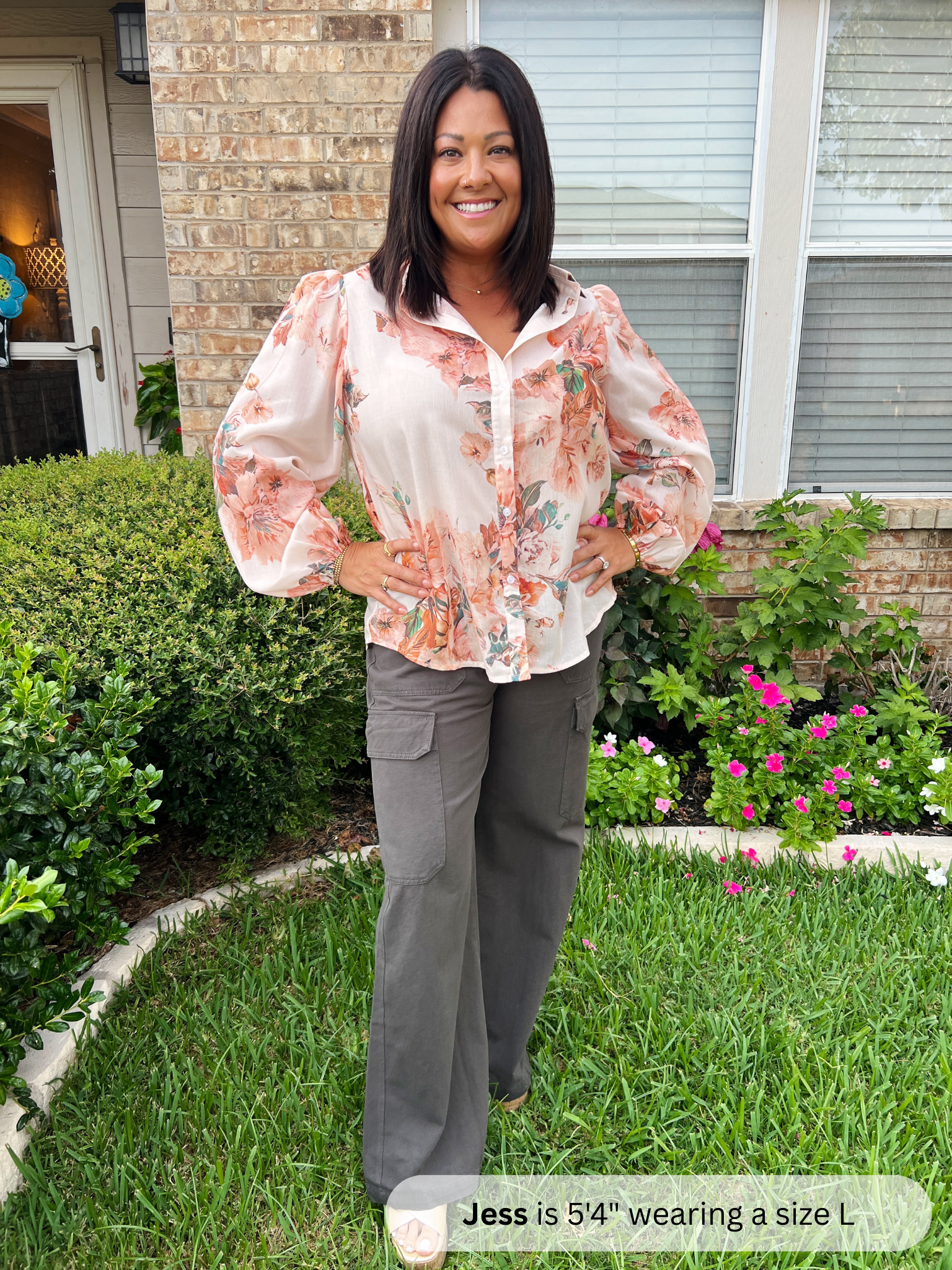 Woman standing outdoors in a garden wearing a floral blouse and gray pants.