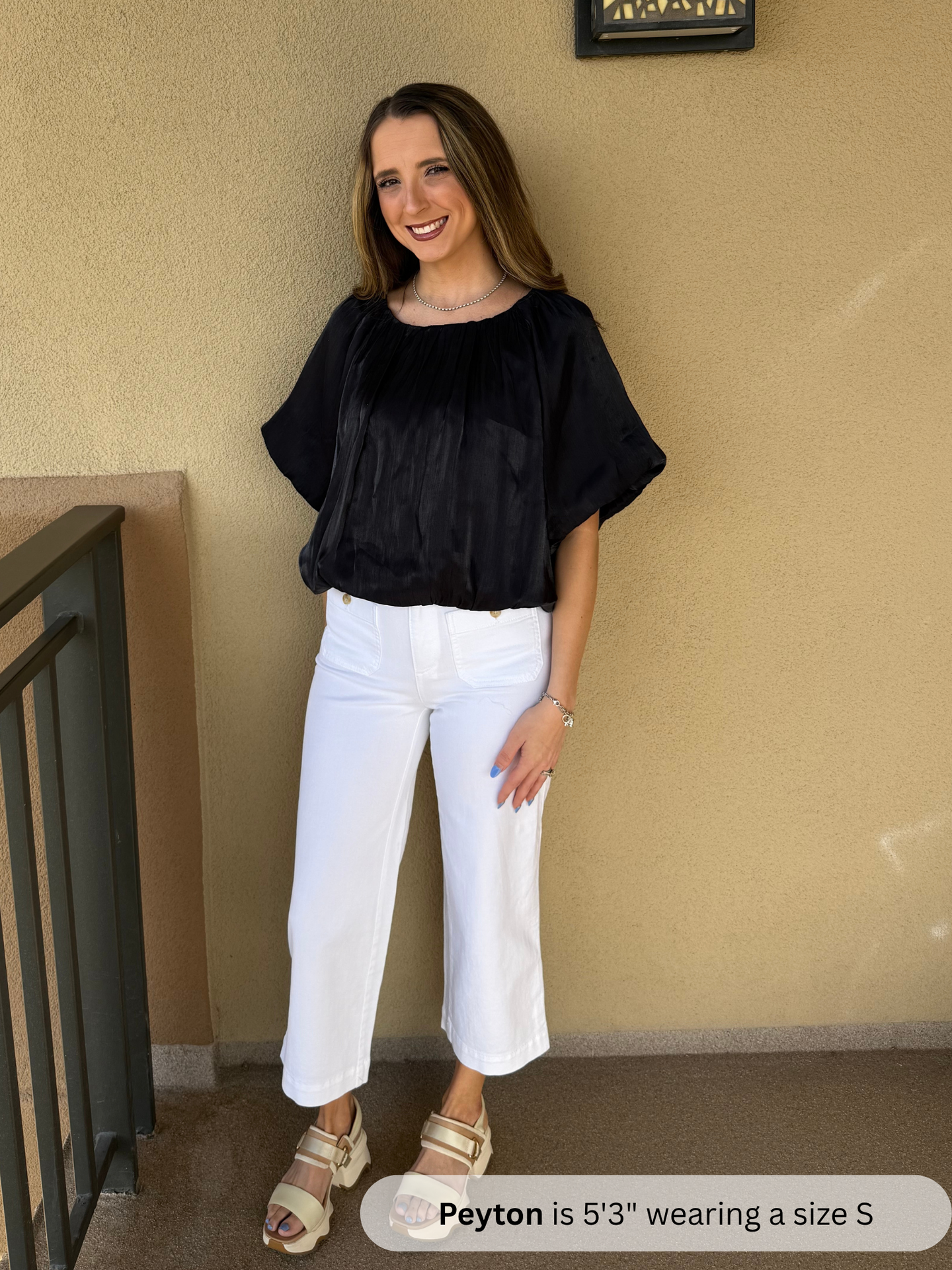 Woman wearing a black top and white pants standing against a beige wall.