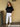Woman wearing a black top and white pants standing against a beige wall.