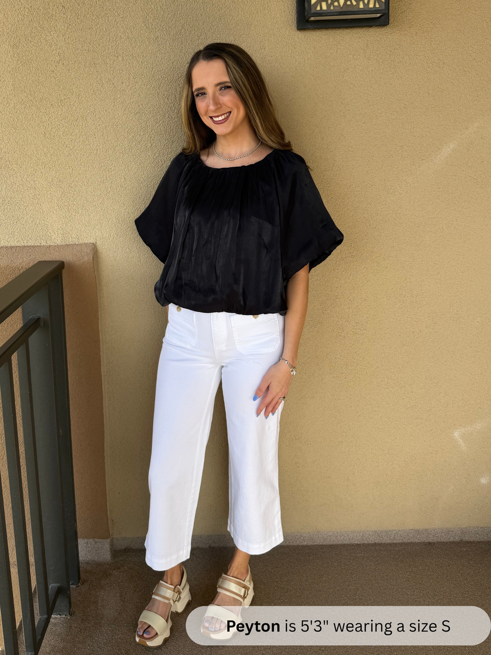 Woman wearing a black top and white pants standing against a beige wall.