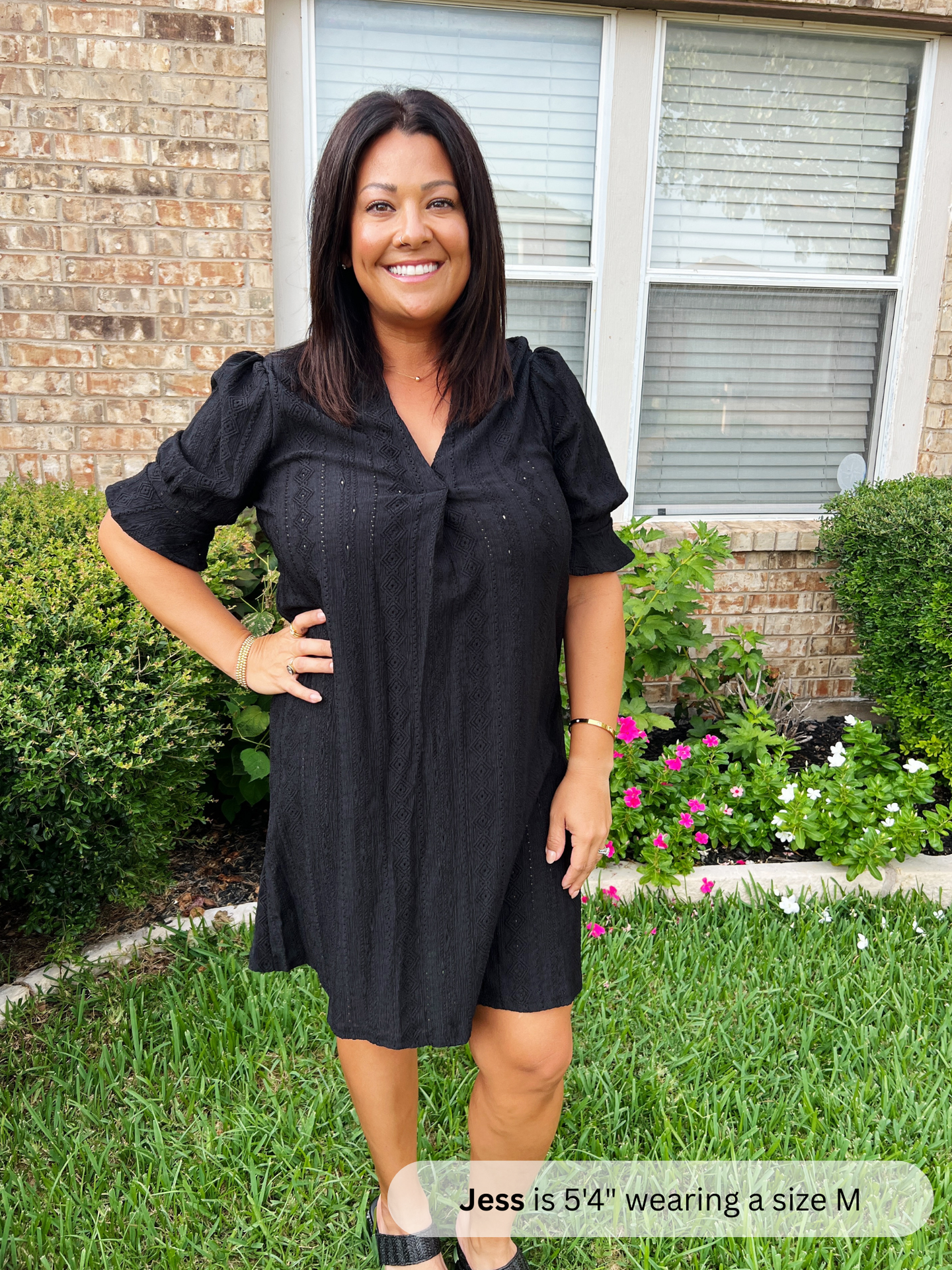 Woman wearing a black dress standing in a garden with a brick wall and window in the background.