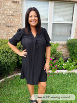 Woman wearing a black dress standing in a garden with a brick wall and window in the background.