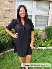 Woman wearing a black dress standing in a garden with a brick wall and window in the background.