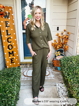 Woman in an olive green outfit standing in front of a house with autumn decorations.