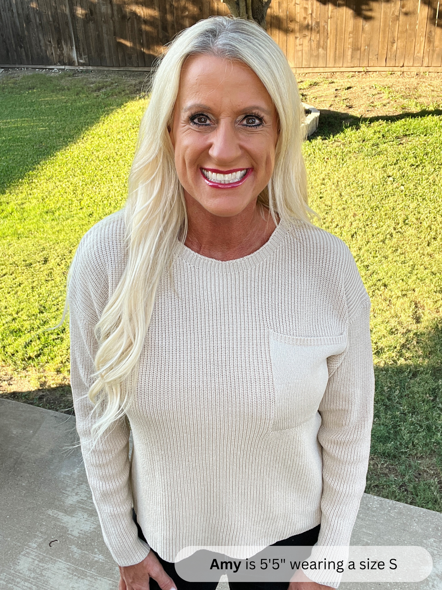 Woman wearing a beige sweater standing outdoors with grass and a wooden fence in the background.