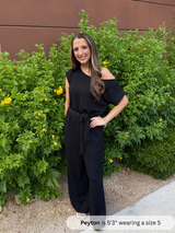 Woman wearing a black off-shoulder jumpsuit standing in front of green bushes.