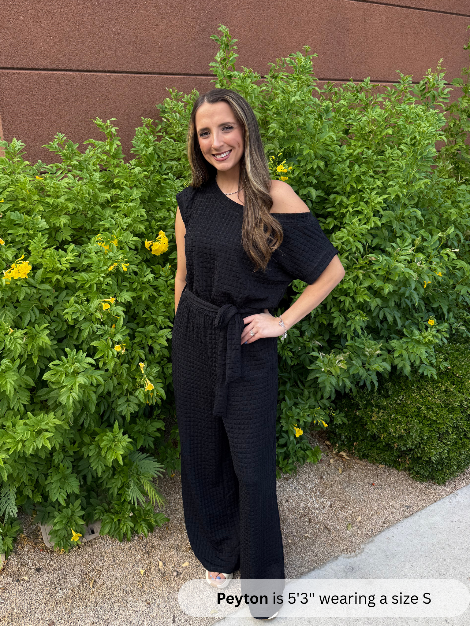Woman wearing a black off-shoulder jumpsuit standing in front of green bushes.
