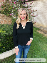 Woman in black top and blue jeans standing outdoors with greenery and a building in the background