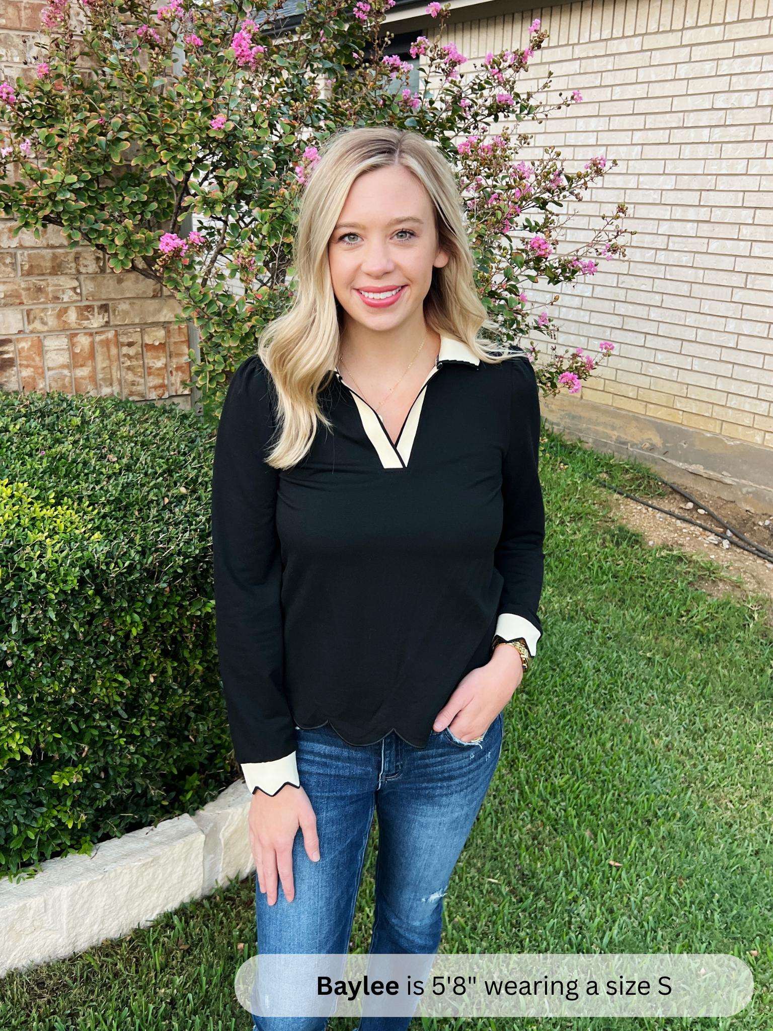 Woman in black top and blue jeans standing outdoors with greenery and a building in the background