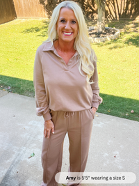 Woman wearing a beige outfit standing on a sidewalk with grass and trees in the background