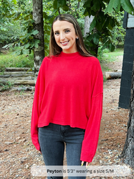 Woman wearing a red sweater and jeans standing outdoors with trees in the background