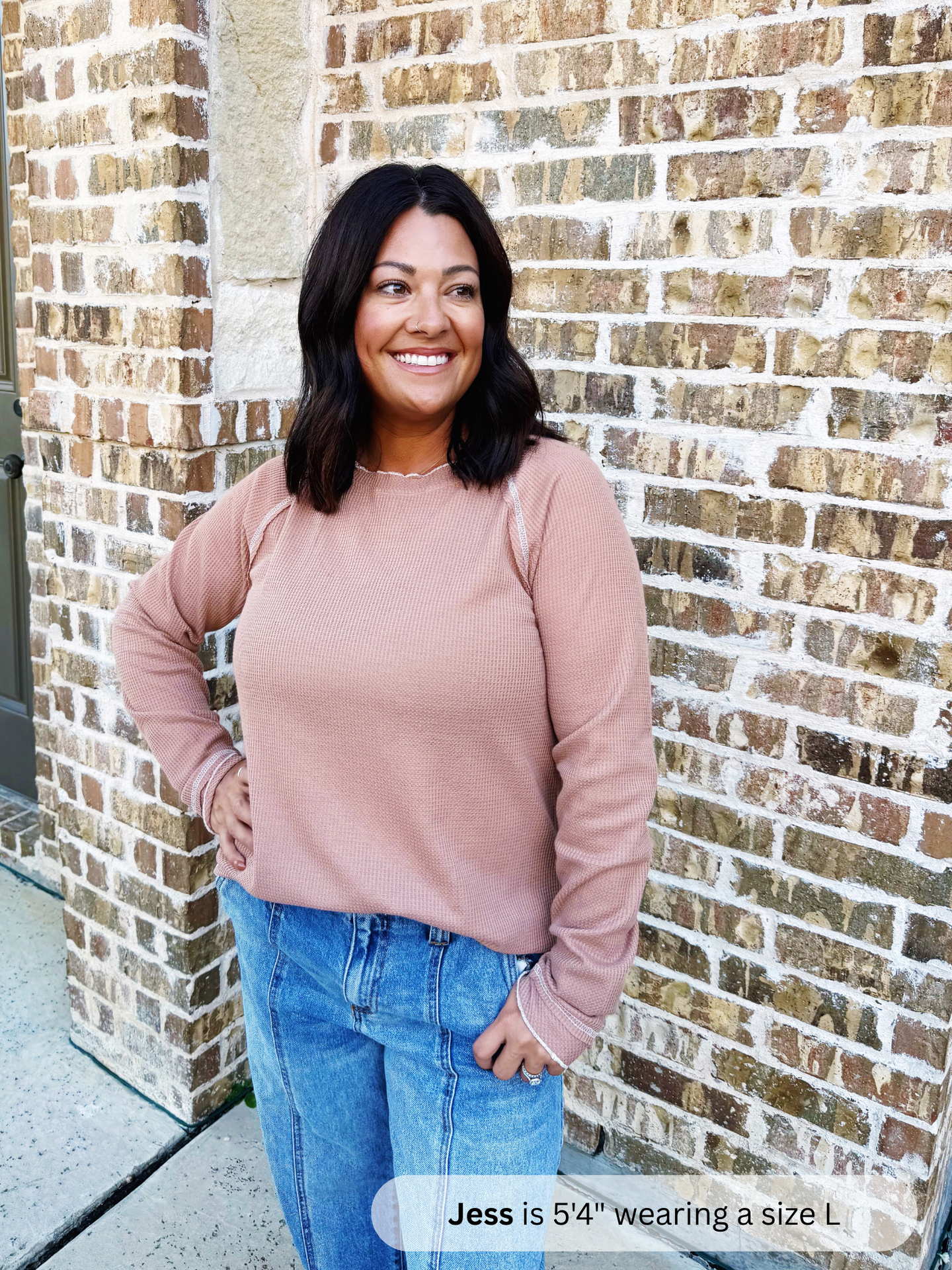 Woman wearing a pink sweatshirt and blue jeans standing against a brick wall.
