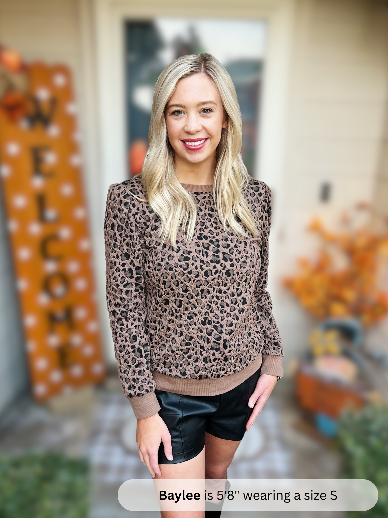 Woman wearing a leopard print sweater and black shorts in an outdoor setting with Halloween decorations.