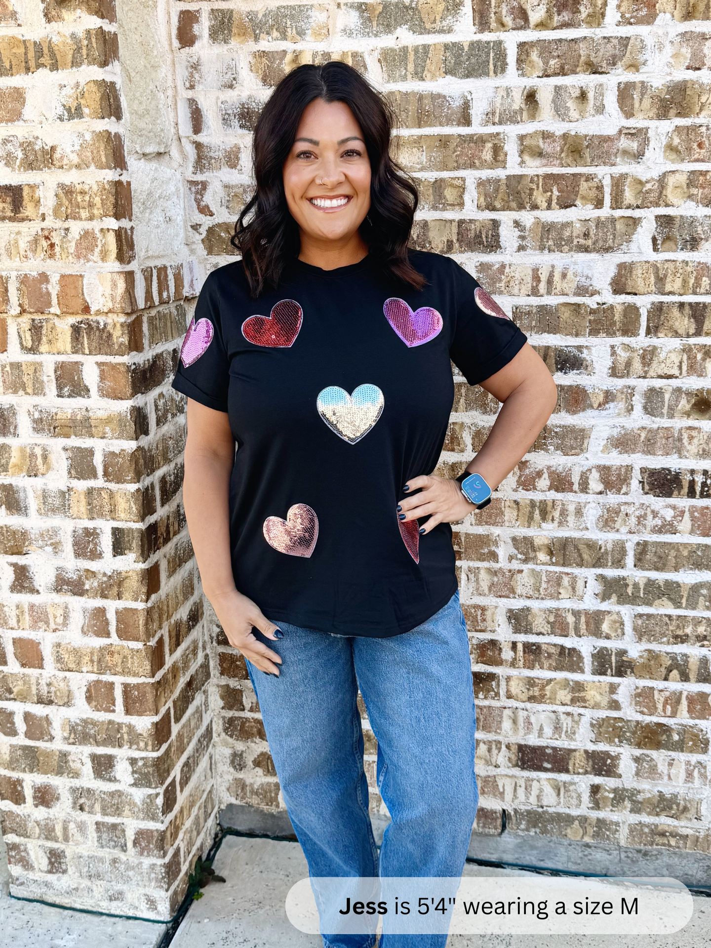 Woman wearing a black t-shirt with colorful heart designs against a brick wall.