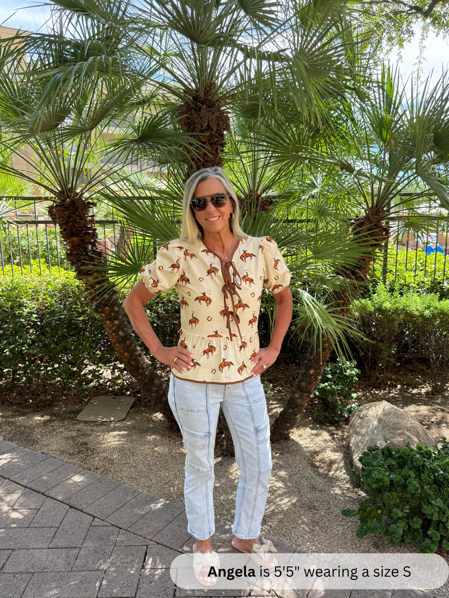 Woman wearing a patterned shirt and white pants standing in front of palm trees.