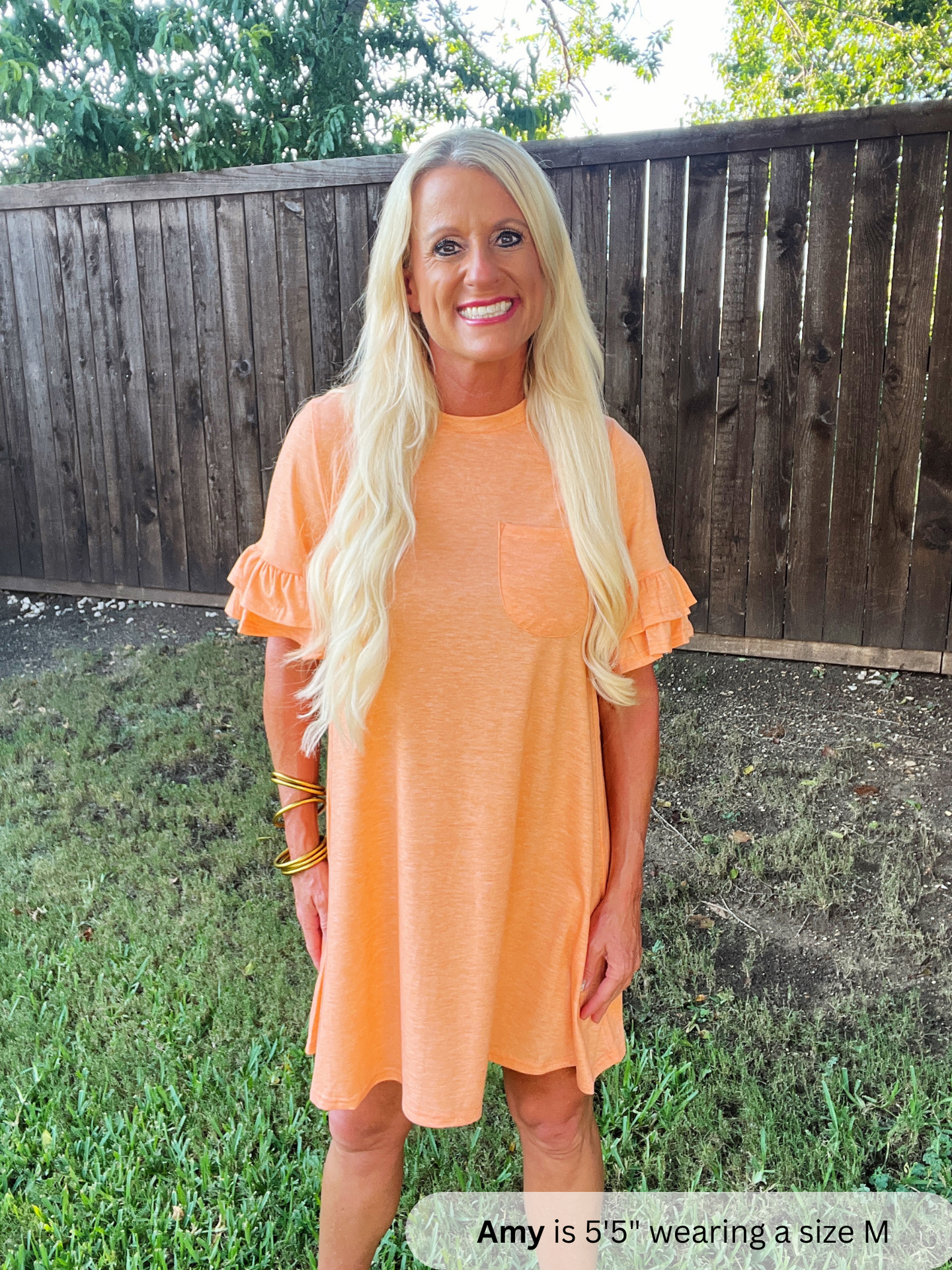 Woman wearing an orange dress standing outdoors with a wooden fence and grass in the background.