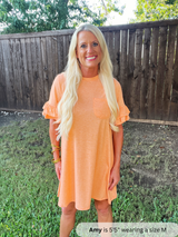 Woman wearing an orange dress standing outdoors with a wooden fence and grass in the background.