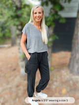Woman wearing a gray top and black pants standing outdoors with trees in the background