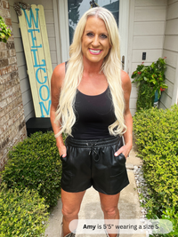 Woman wearing a black tank top and leather shorts standing in front of a house with a 'Welcome' sign.