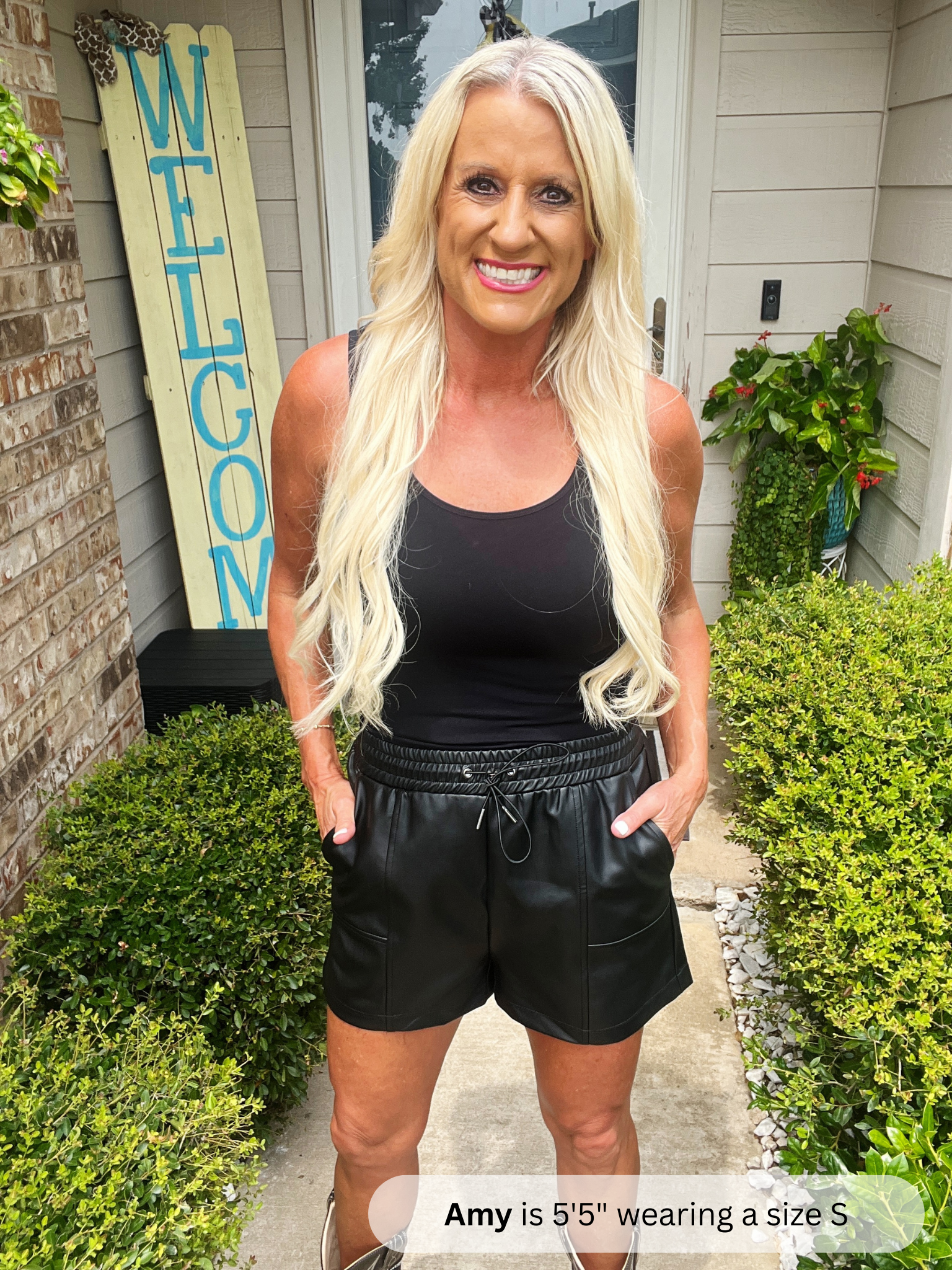 Woman wearing a black tank top and leather shorts standing in front of a house with a 'Welcome' sign.
