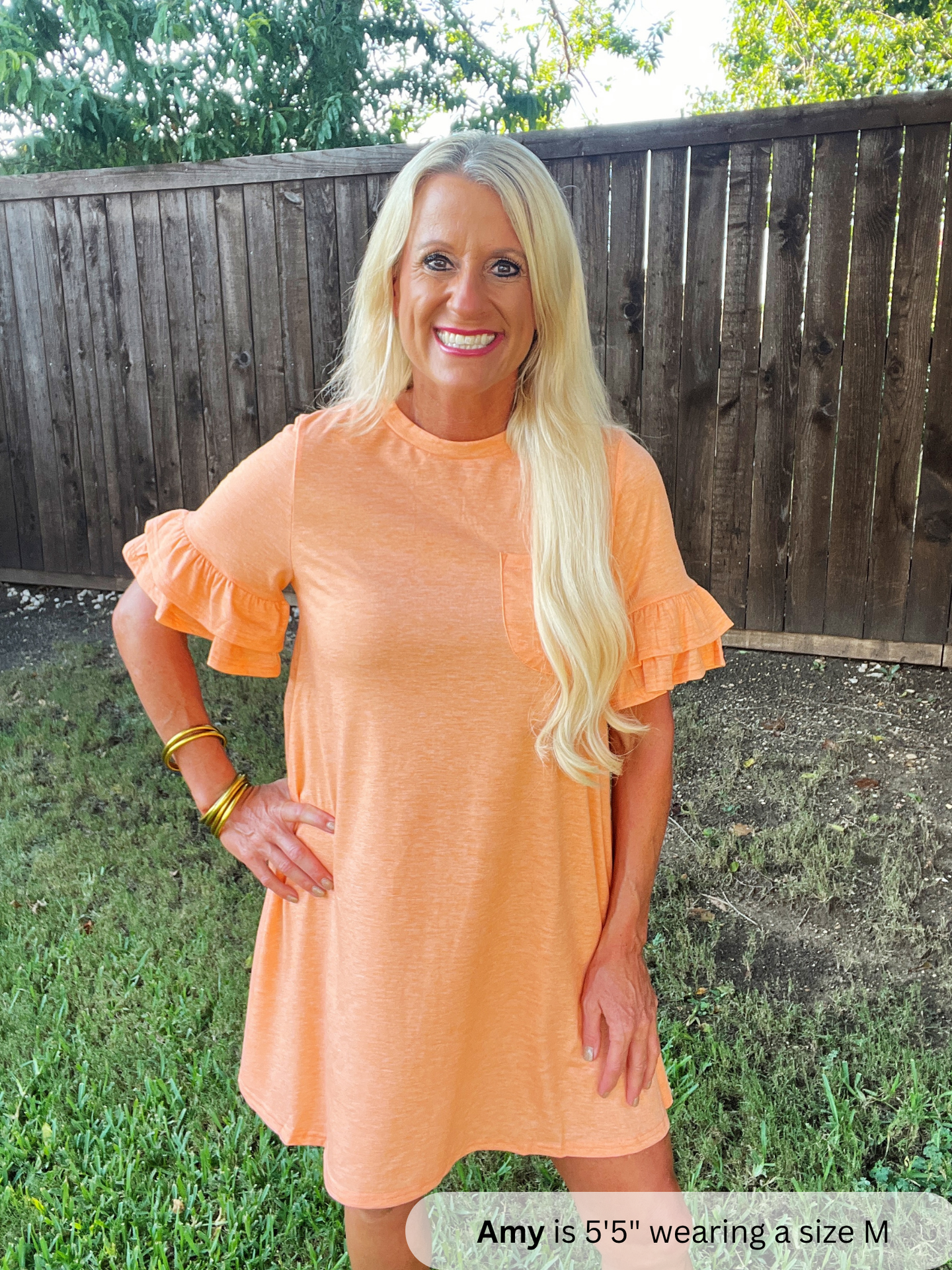 Woman wearing an orange dress standing outdoors with a wooden fence and grass in the background.
