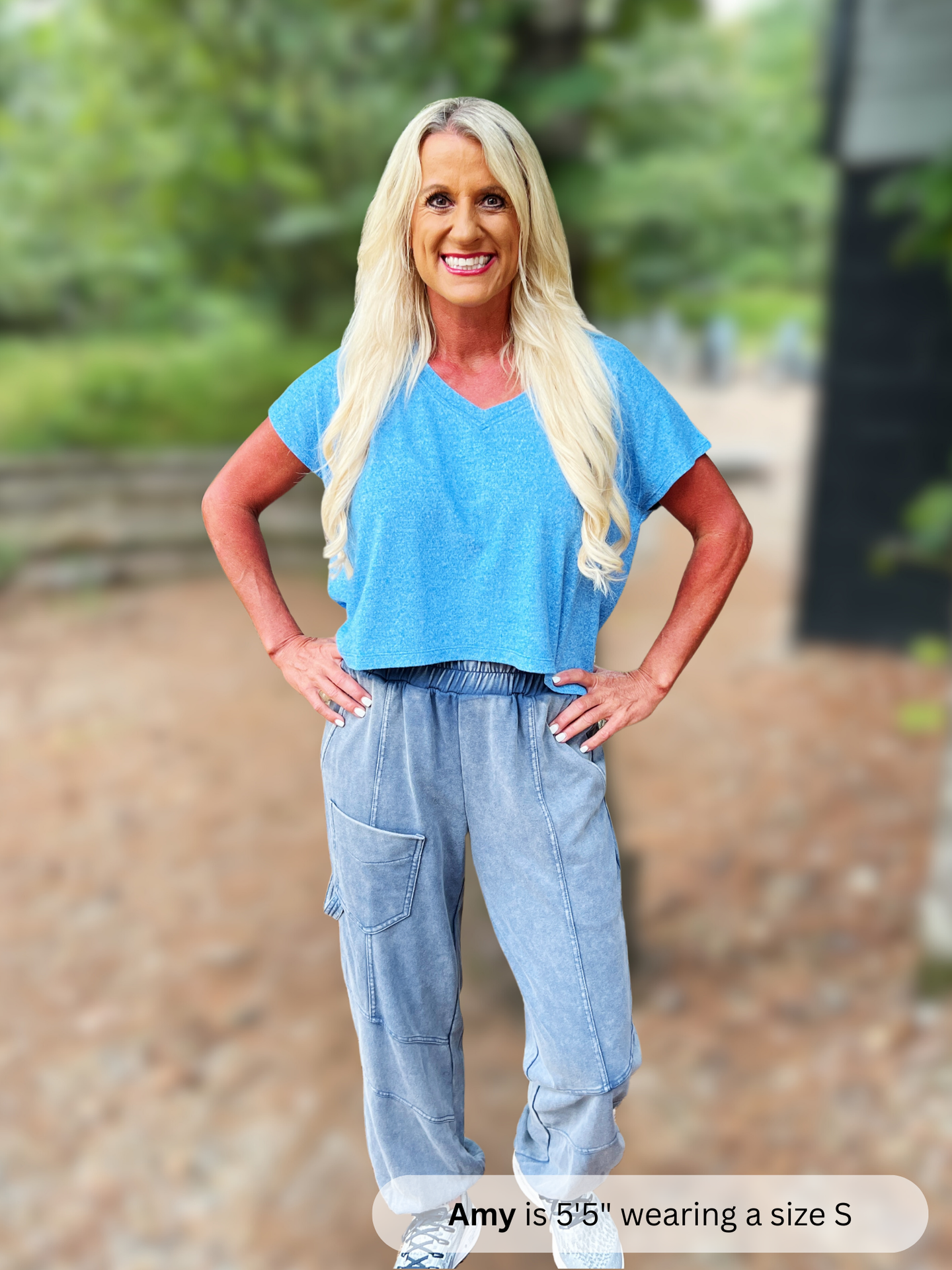 Woman wearing a blue shirt and light blue pants standing outdoors with trees in the background