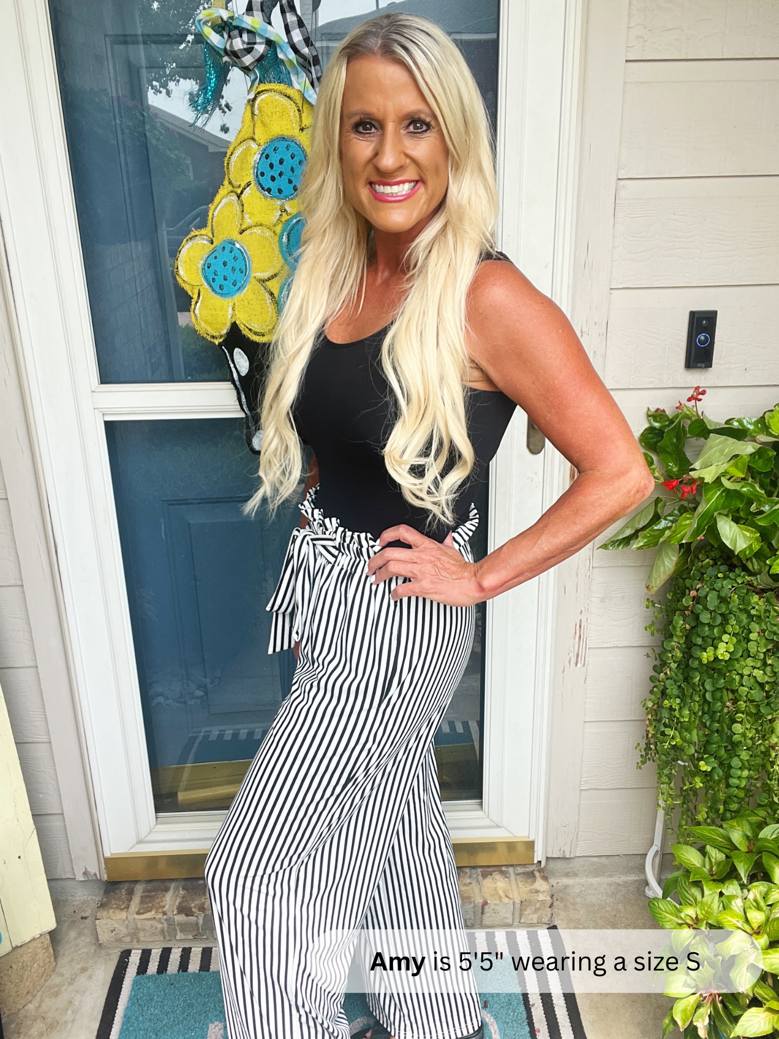 Woman standing in front of a door with a decorative flower, wearing a black top and striped pants.