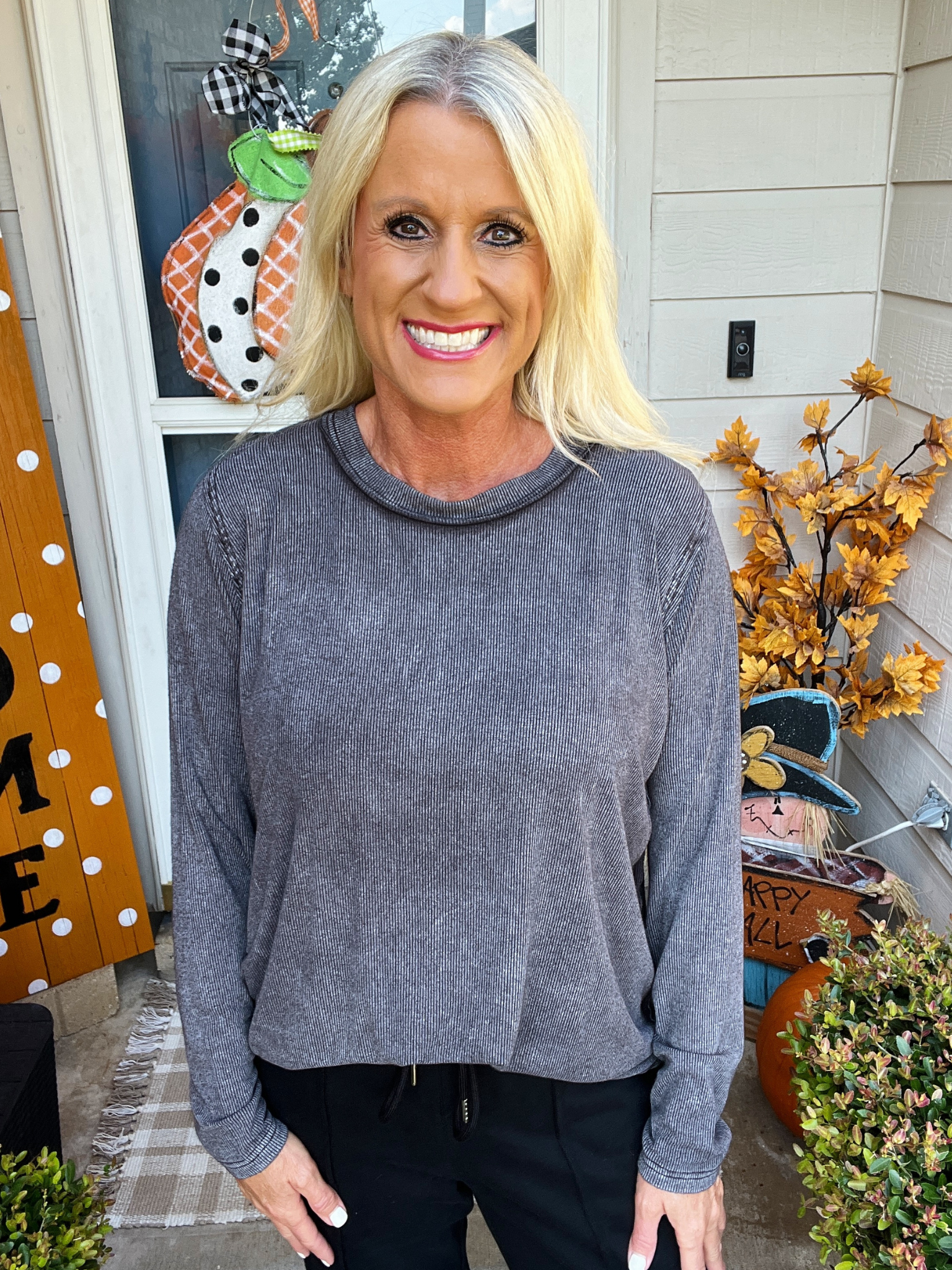Woman wearing a gray sweater standing in front of a decorated door with pumpkins and autumn decorations.