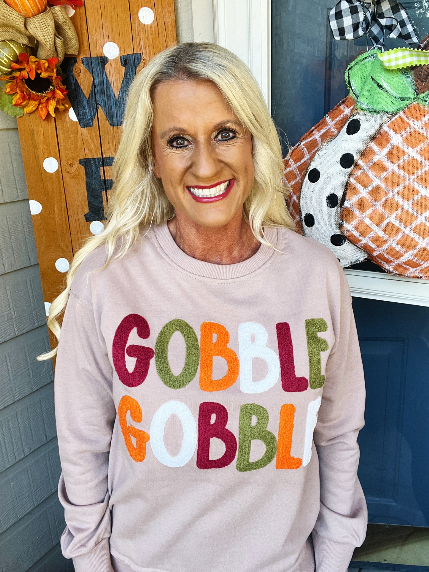 Woman wearing a 'Gobble Gobble' shirt in front of a decorated door with pumpkins and autumn decorations.