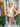 Woman standing in front of a house with a 'Welcome' sign, wearing a beige top and brown skirt.