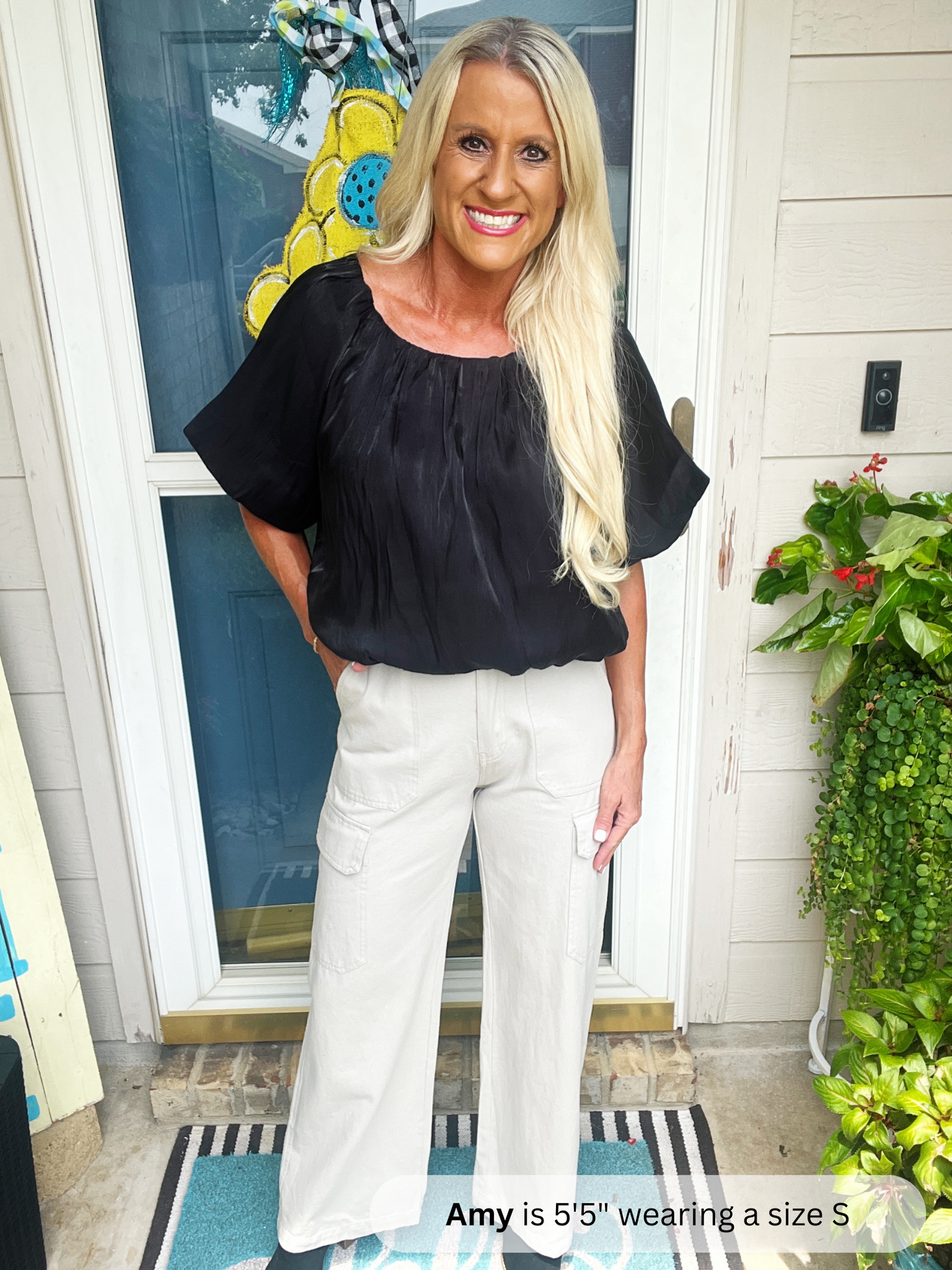 Woman wearing a black top and white pants standing in front of a door with plants around.