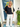 Woman wearing a black top and white pants standing in front of a door with plants around.