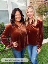 Two women wearing brown velvet tops standing outdoors with greenery and a house in the background.