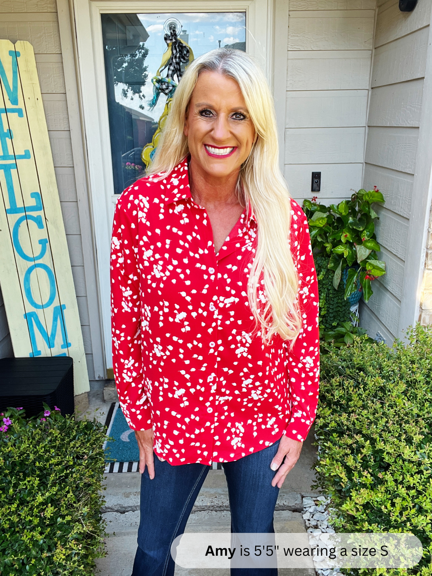 Woman wearing a red floral blouse standing outside a house with 'WELCOME' sign.