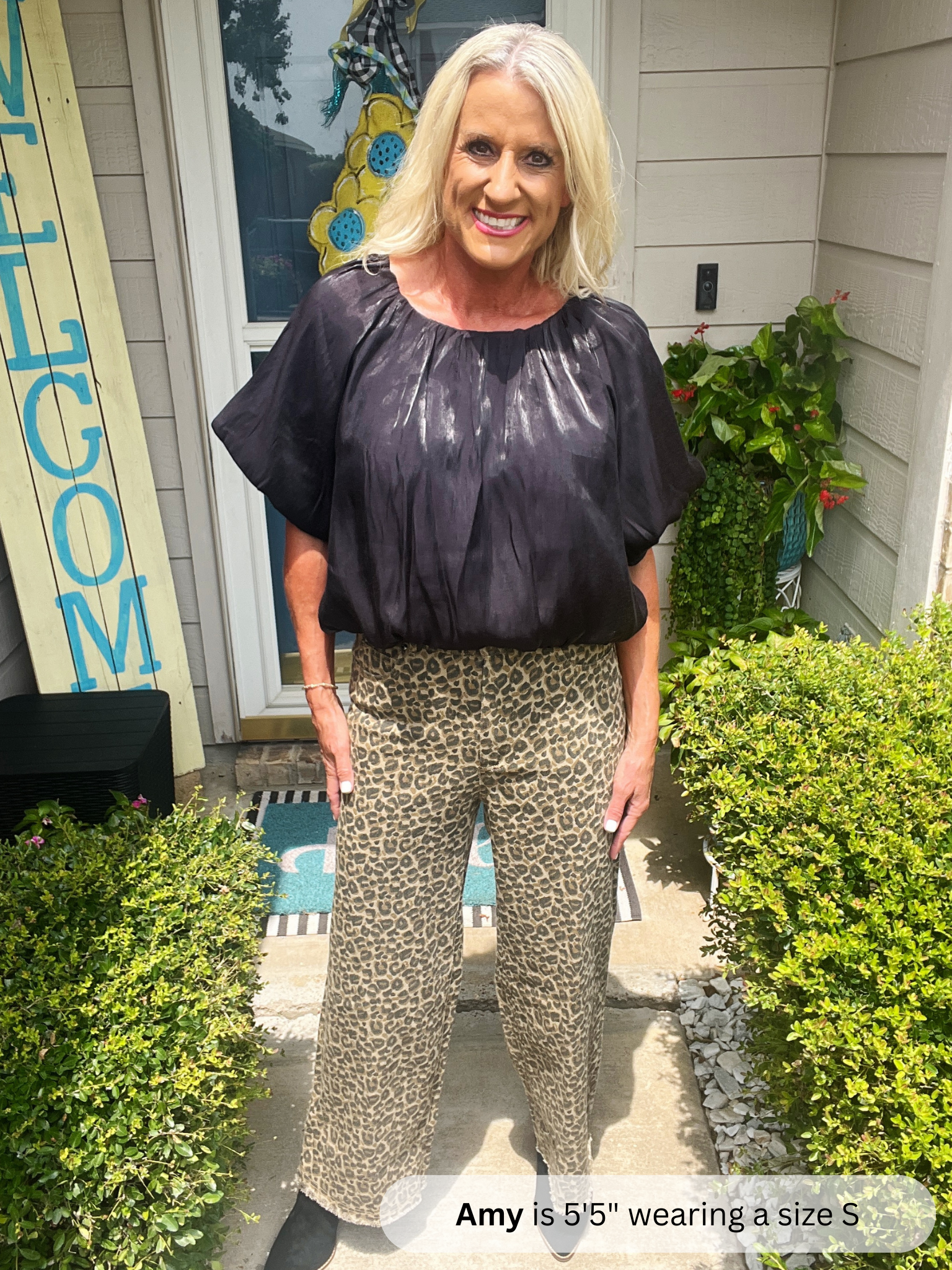 Woman standing outside a house wearing a black top and leopard print pants.