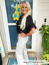 Woman standing outside a house with a decorative wreath, wearing a black top and white pants.
