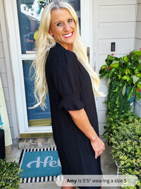 Woman wearing a black dress standing outside a house with plants and a 'Hello' mat.
