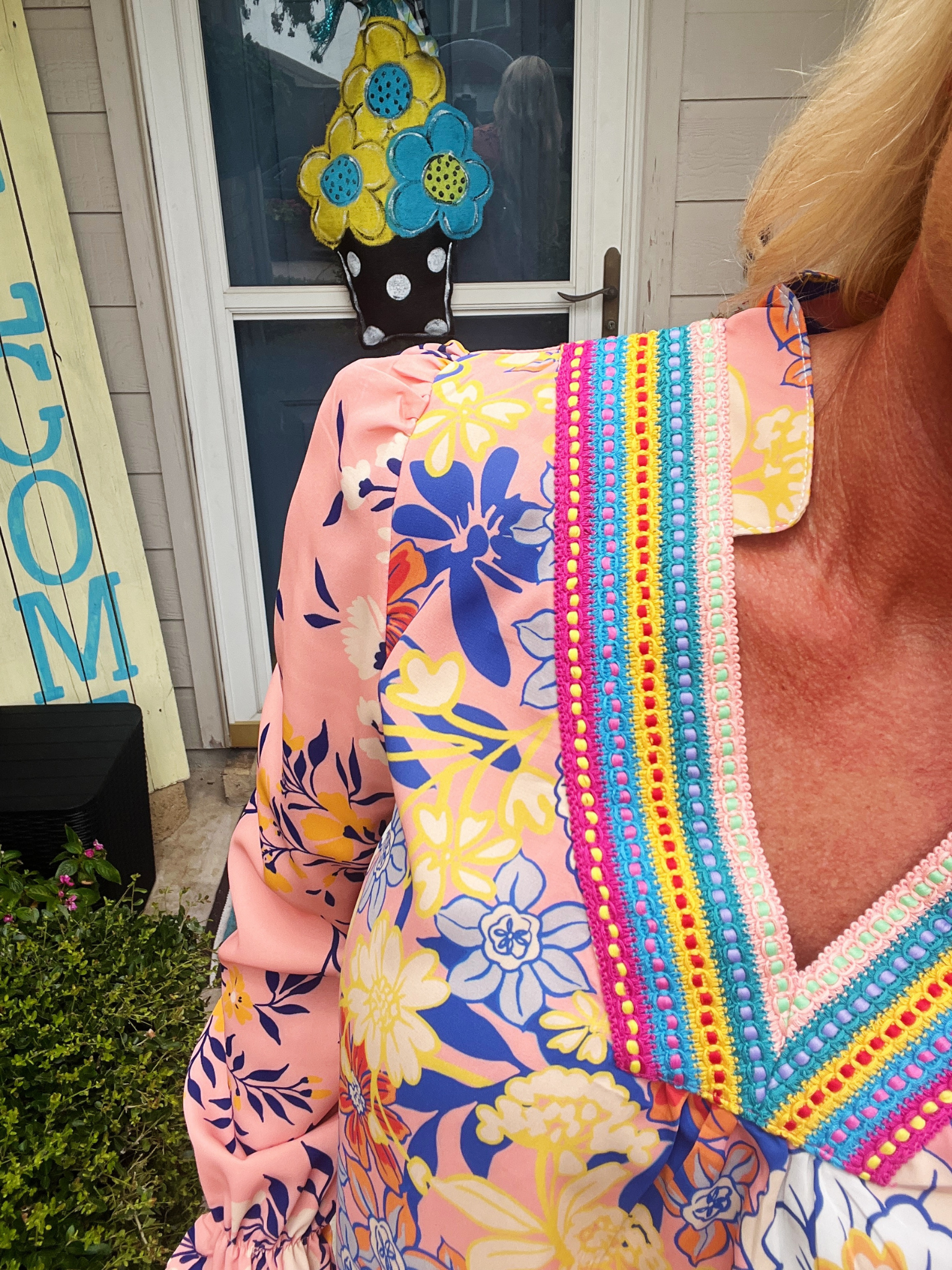 Person wearing a colorful floral dress with a vibrant trim, standing in front of a 'Welcome' sign.