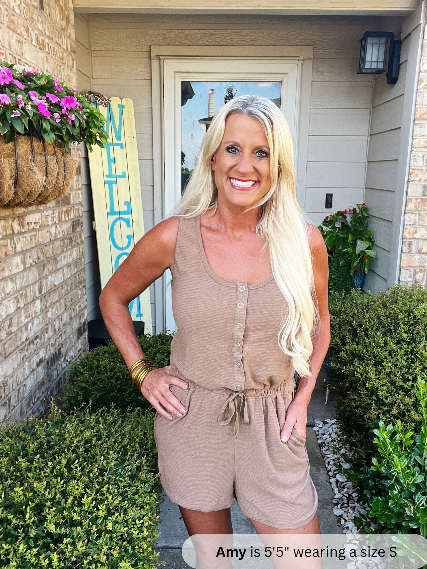 /Mocha/ Woman in a beige romper standing outside a house with plants and a surfboard in the background.