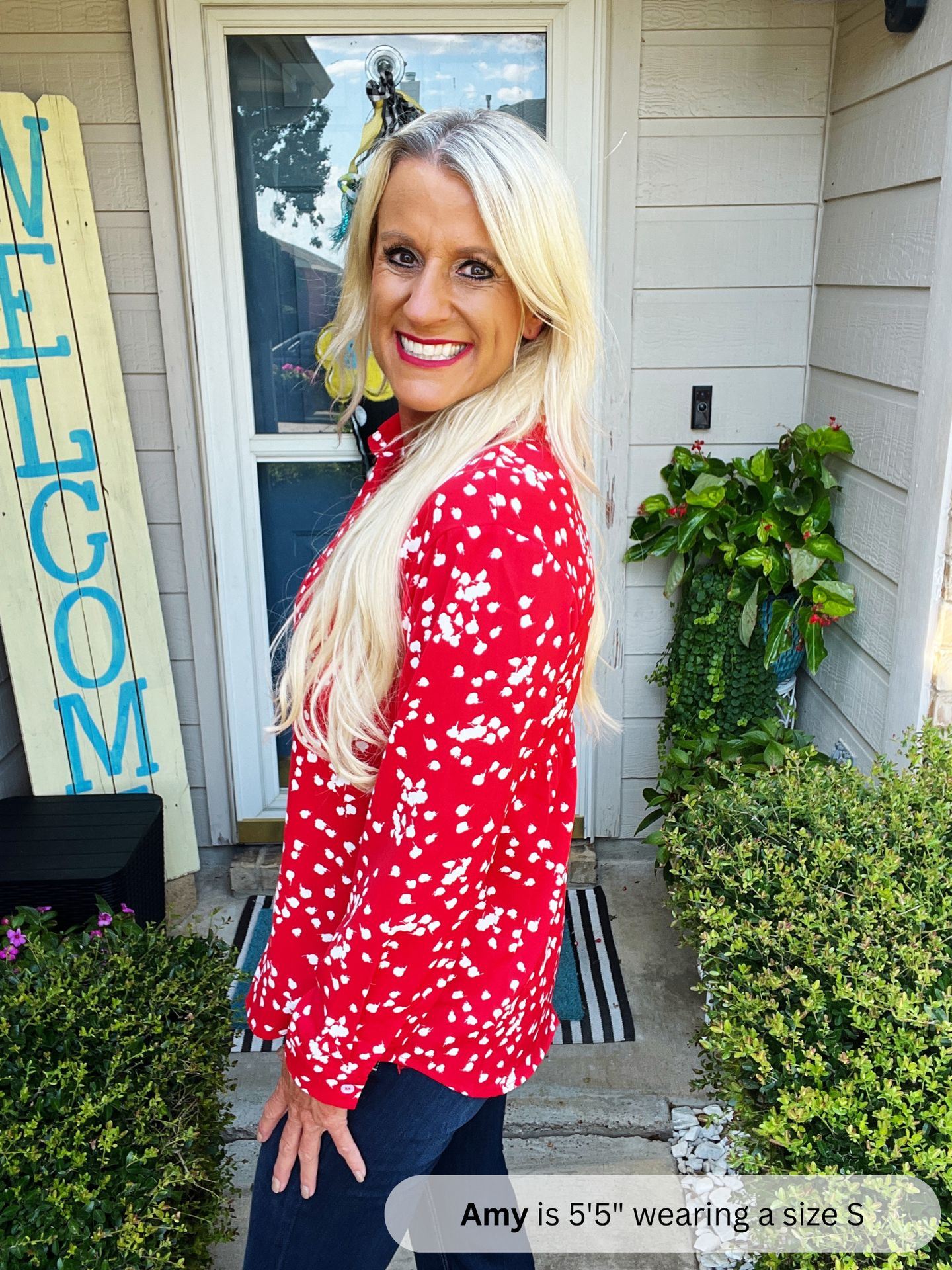 Woman in a red floral blouse standing outside a house with a 'WELCOME' sign.