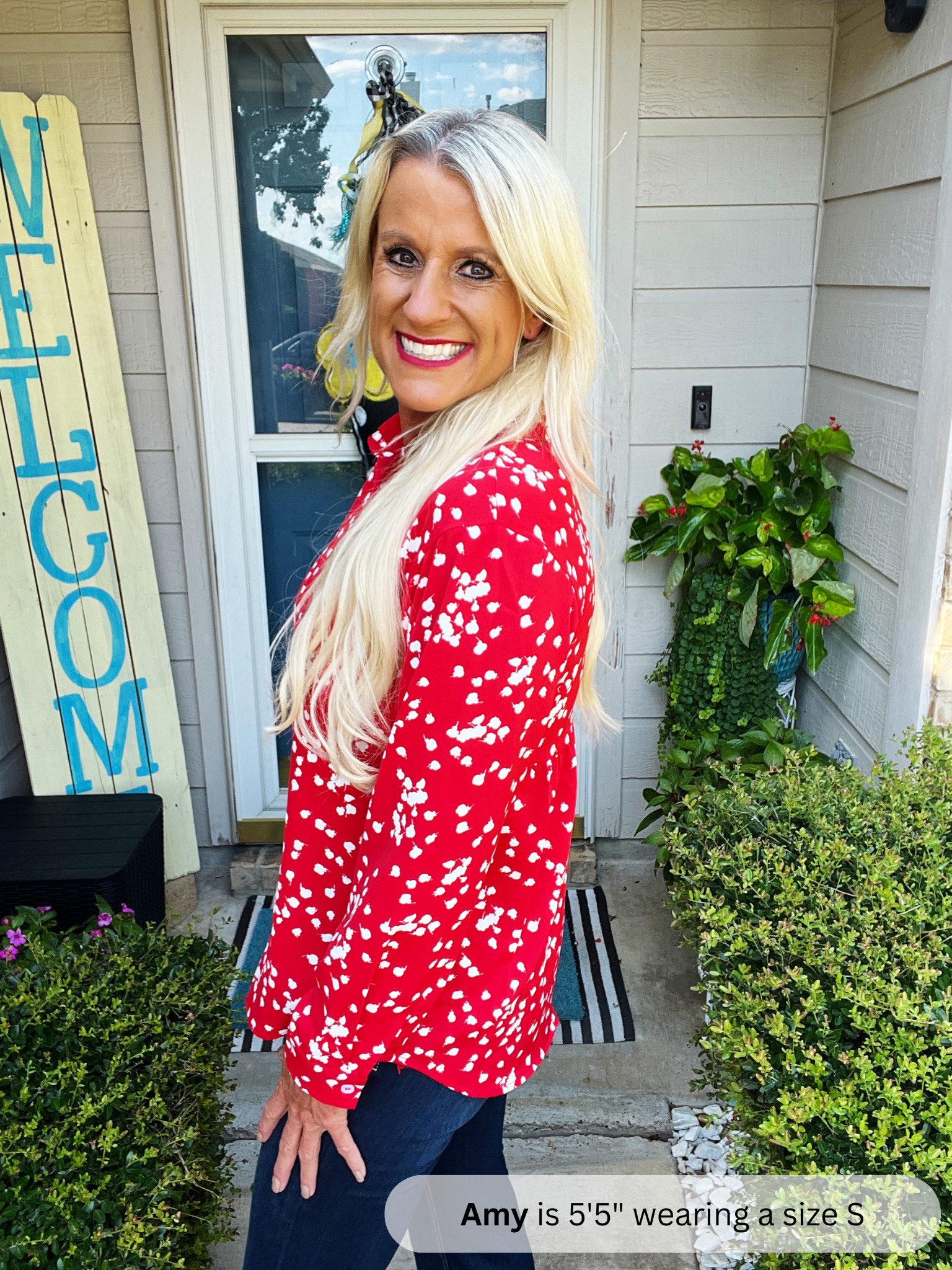 Woman in a red floral blouse standing outside a house with a 'WELCOME' sign.